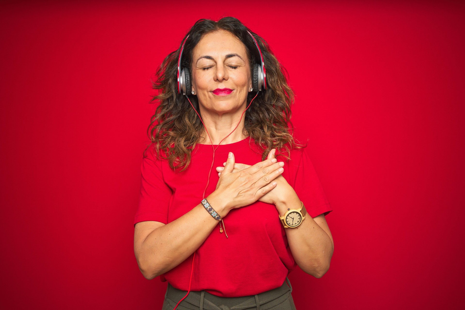 Middle age senior woman wearing headphones listening to music over red isolated background smiling with hands on chest with closed eyes and grateful gesture on face. Health concept.